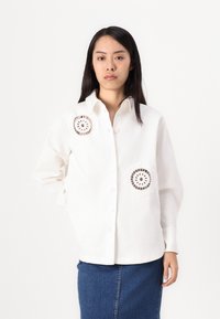 Cămașă albă cu nasturi, cu detalii brodate rotunde pe buzunare, mâneci lungi și guler clasic. Asortată cu o fustă din denim.