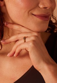 Gouden gedraaide ring met een gladde oppervlakte, gedragen aan een vinger, met een close-up van hand en gezicht, tegen een neutrale achtergrond.