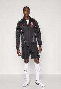 Sort atletisk jakke med røde accenter og hvide striber, parret med sorte shorts, hvide sokker og fodboldstøvler. Bemærk logoernes placering.