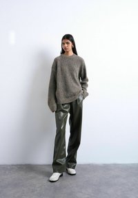 Femme debout contre un mur blanc, portant un pull gris oversize, un pantalon en cuir vert foncé, des chaussures blanches et des boucles d'oreilles en or, les mains dans les poches.