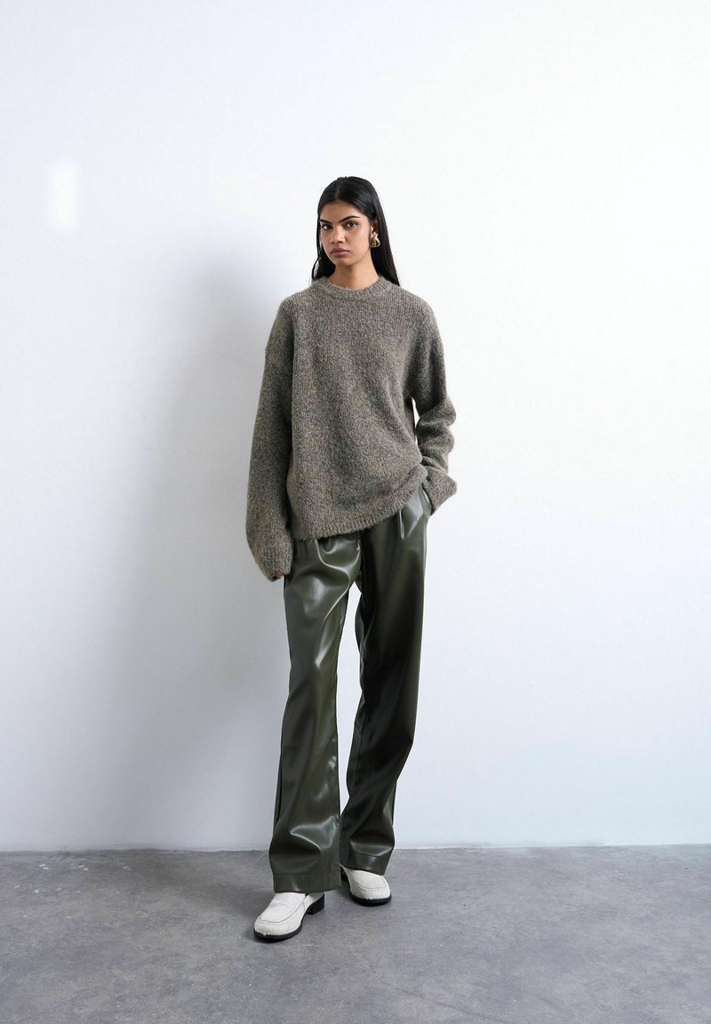 Femme debout contre un mur blanc, portant un pull gris oversize, un pantalon en cuir vert foncé, des chaussures blanches et des boucles d'oreilles en or, les mains dans les poches.