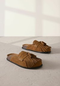 Zapatos de ante marrón tipo slip-on con detalle de flecos, suela de goma plana y un acento cosido. Textura suave y diseño casual.