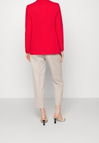 Persona in piedi, di spalle, che indossa un blazer rosso brillante, pantaloni beige a vita alta e sandali beige con tacco su uno sfondo uniforme.