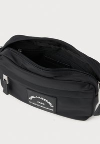 Melnas neilona soma ar taisnstūra formu, ar priekšējo logo uzlīmi, ar rāvējslēdzēju galveno nodalījumu un satīna apdari; ar regulējamu siksnu.