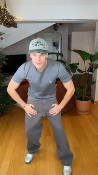 Graues T-Shirt, graue Hose und silberne Sneaker. Die Person posiert mit den Händen auf den Knien. Innenraum mit einer Pflanze und einem Sofa im Hintergrund.