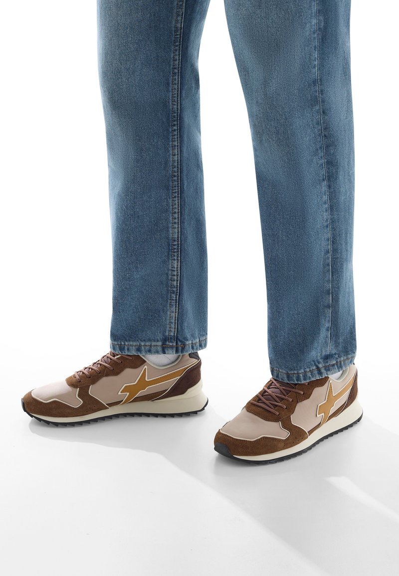 Persona che indossa jeans blu e sneaker atletiche marrone-beige con suole bianche, in piedi su una superficie bianca.