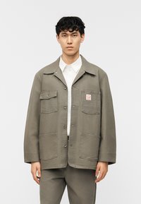 Νέος άνδρας όρθιος, φορώντας πράσινο μπουφάν με κουμπιά και τέσσερις μπροστινές τσέπες πάνω από ένα λευκό πουκάμισο με γιακά και αντίστοιχο παντελόνι.