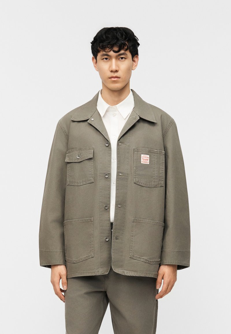 Νέος άνδρας όρθιος, φορώντας πράσινο μπουφάν με κουμπιά και τέσσερις μπροστινές τσέπες πάνω από ένα λευκό πουκάμισο με γιακά και αντίστοιχο παντελόνι.