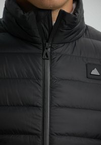 Close-up van een zwart gewatteerd jack, tot de nek dichtgeritst, met een rubberen patch met een berglogo op de borst.