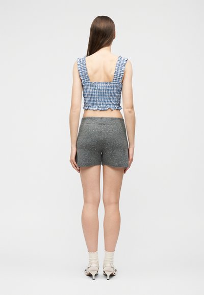 Femme debout de dos, portant un crop top sans manches à carreaux bleus, un short taille haute gris avec de petits pois, des chaussettes blanches et des talons.