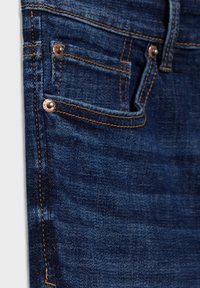 Pantalones vaqueros de denim azul oscuro con costuras visibles, acentos de hardware en cobre, incluidos remaches, y una textura ligeramente desgastada cerca del bolsillo frontal.