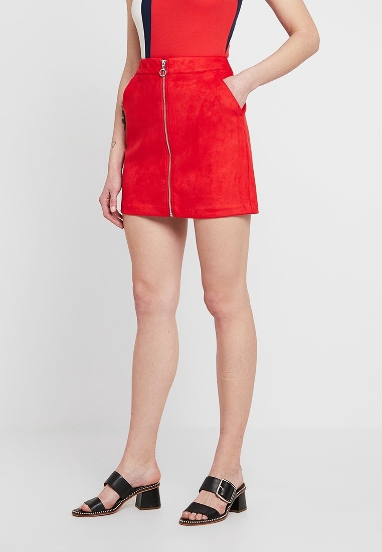 Mini-jupe en daim rouge avec fermeture éclair à l'avant, deux poches latérales et une coupe ajustée. Associée à des mules à talons blocs noires.