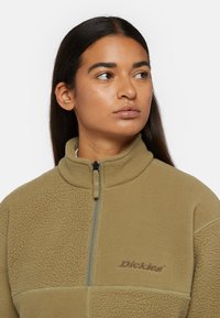 Chaqueta de forro polar con cremallera de cuarto en verde oliva, con un alto collar, tejido texturizado y logo bordado en el pecho. Detalles de cremallera visibles.