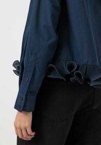 Marineblauwe blouse met een mouw met knoopsleave en een gerimpelde zoom, gedragen met zwarte denim jeans aan de linkerkant van een persoon.