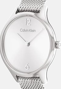 Calvin Klein hõbe kell, metallvõrgust rihm, ümmargune korpus, valge sageduse dial, minimalistlikud tundemärgid 12 ja 6, sujuv tekstuur, klassikaline disain.