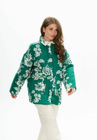 Groene gewatteerde jas met witte bloemmotieven, voorzien van een knoopsluiting, grote zakken en een contrasterende witte kraag.