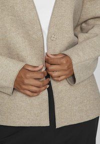 Beige ullblandad jacka med en enda rund knappstängning, vida ärmar och råa kanter, tillsammans med en vit topp och svarta byxor.