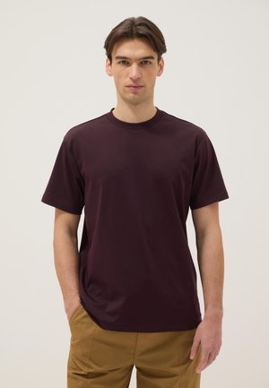 Jeune homme aux cheveux bruns courts portant un t-shirt bordeaux foncé à manches courtes et un pantalon beige, debout devant un fond clair.