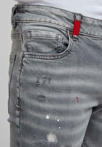 Szürke, kopott farmernadrág festékfoltokkal és egy piros címkével a derékrészen, fehér, derék fölött vágott felsővel viselve.