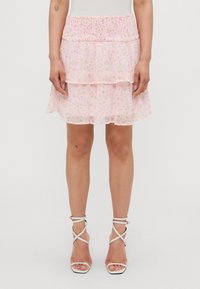 Jupe rose à couches avec motif floral, taille élastique, tissu transparent et ourlet volanté. Portée avec des sandales à talons blancs à lanières.