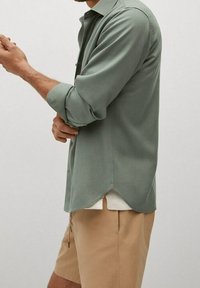 Lichtgroen overhemd met lange mouwen. Heeft een gebogen zoom en een gladde textuur, gedragen over een wit T-shirt en beige shorts.