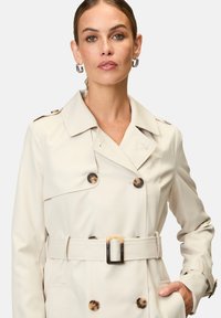 Beige Trenchcoat mit Gürtel, ausgestattet mit großen braunen Knöpfen, einem doppelreihigen Design und Schulterklappen. Glatte Stofftextur.