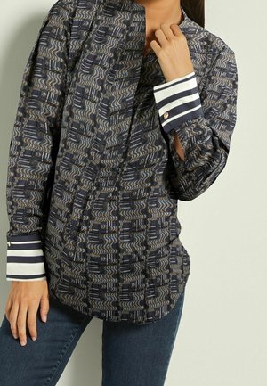 Femme portant une chemise noire à manches longues et motifs, avec des poignets rayés blancs et noirs, associée à un jean bleu, main près de son col.