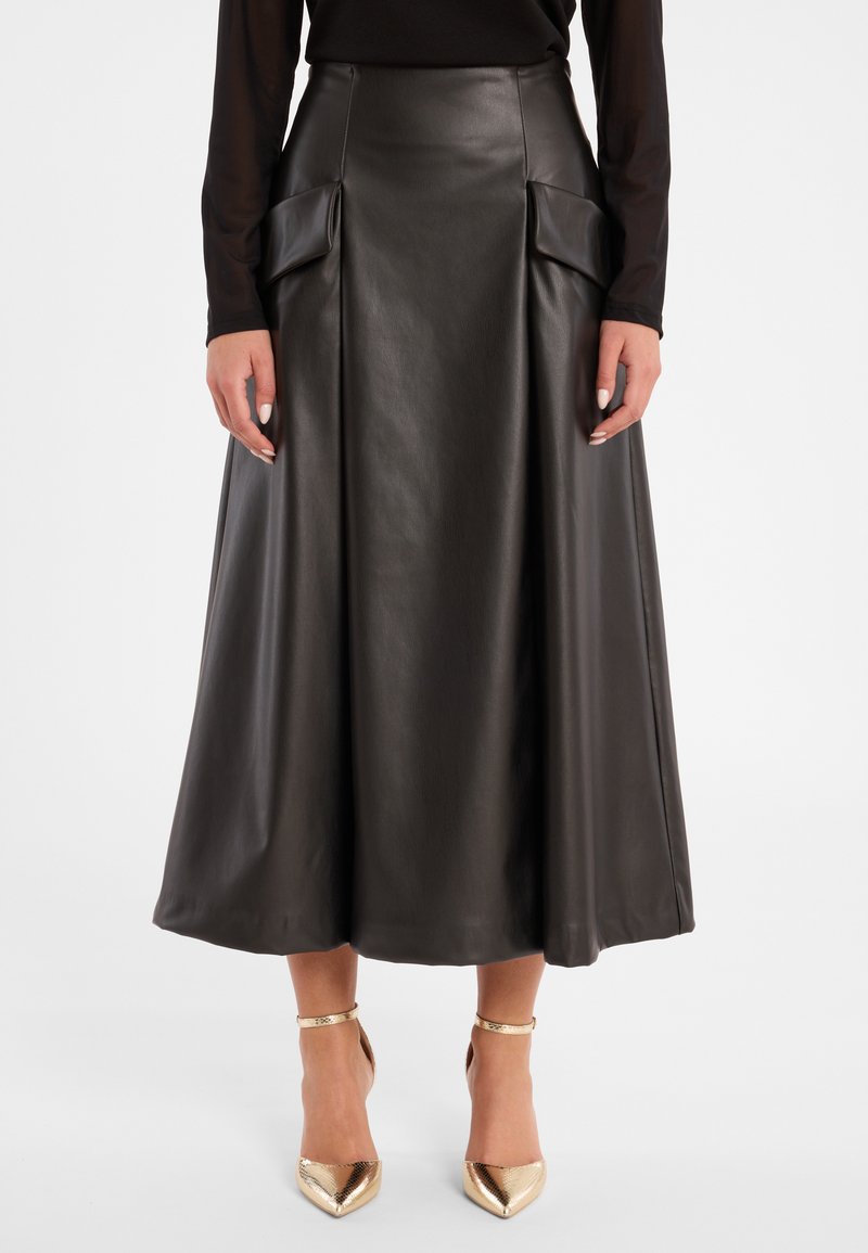 Schwarzer Leder-Midikleid mit hohem Bund, vorderen Falten und zwei großen Taschen. Kombiniert mit glänzenden goldenen spitzen High Heels.