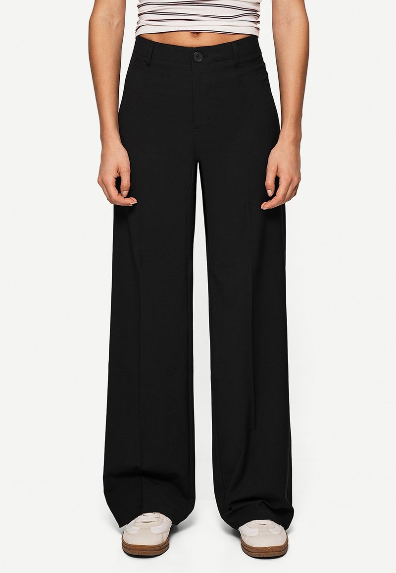 Personne portant un pantalon noir ample, des baskets blanches et un top court blanc rayé noir, debout devant un fond uni.
