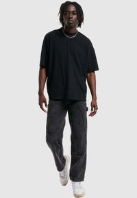 Black oversized t-shirt with short sleeves, paired with dark jeans featuring pockets and a textured finish. Worn with white sneakers and chain.