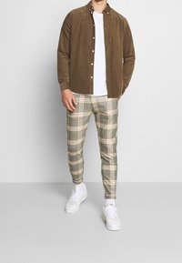 Man i brun knappad skjorta över vit t-shirt, grå-och-beige rutiga byxor och vita sneakers som står framför en enkel bakgrund.