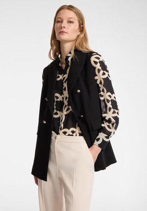Gilet nero su misura con bottoni doppi, indossato sopra una blusa a fantasia crema e nera; i pantaloni crema completano l'outfit.