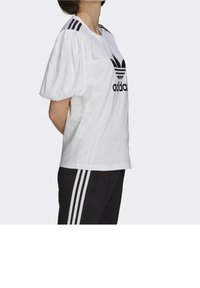 Camiseta blanca con mangas abullonadas, logo negro de adidas en el frente y tres rayas negras en los hombros. Se lleva con pantalones negros con rayas.