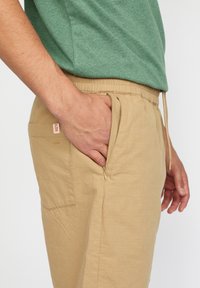 Khaki lichtgewicht broek met een elastische tailleband, zijzakken en een ritszak, en een gestructureerde stof. Groene top gedeeltelijk zichtbaar.