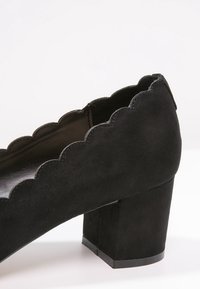 Black suede high-heeled shoe with a chunky block heel and scalloped edge detailing. Textured surface with a smooth interior lining.