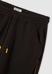 Pantaloni tuta neri con vita elastica, stringhe nere con punte gialle, tasche laterali e una piccola etichetta bianca del marchio all'interno della vita.