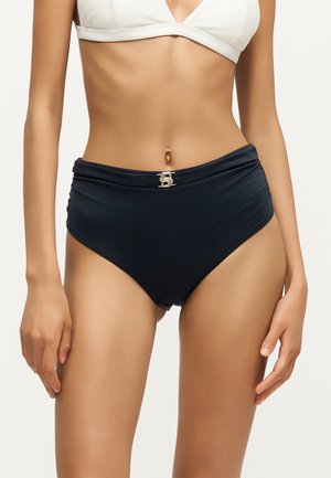 Kvinde iført sort bikini underdel med høj talje og hvid bikini top, der viser et gyldent dekorativt spænde på taljen.