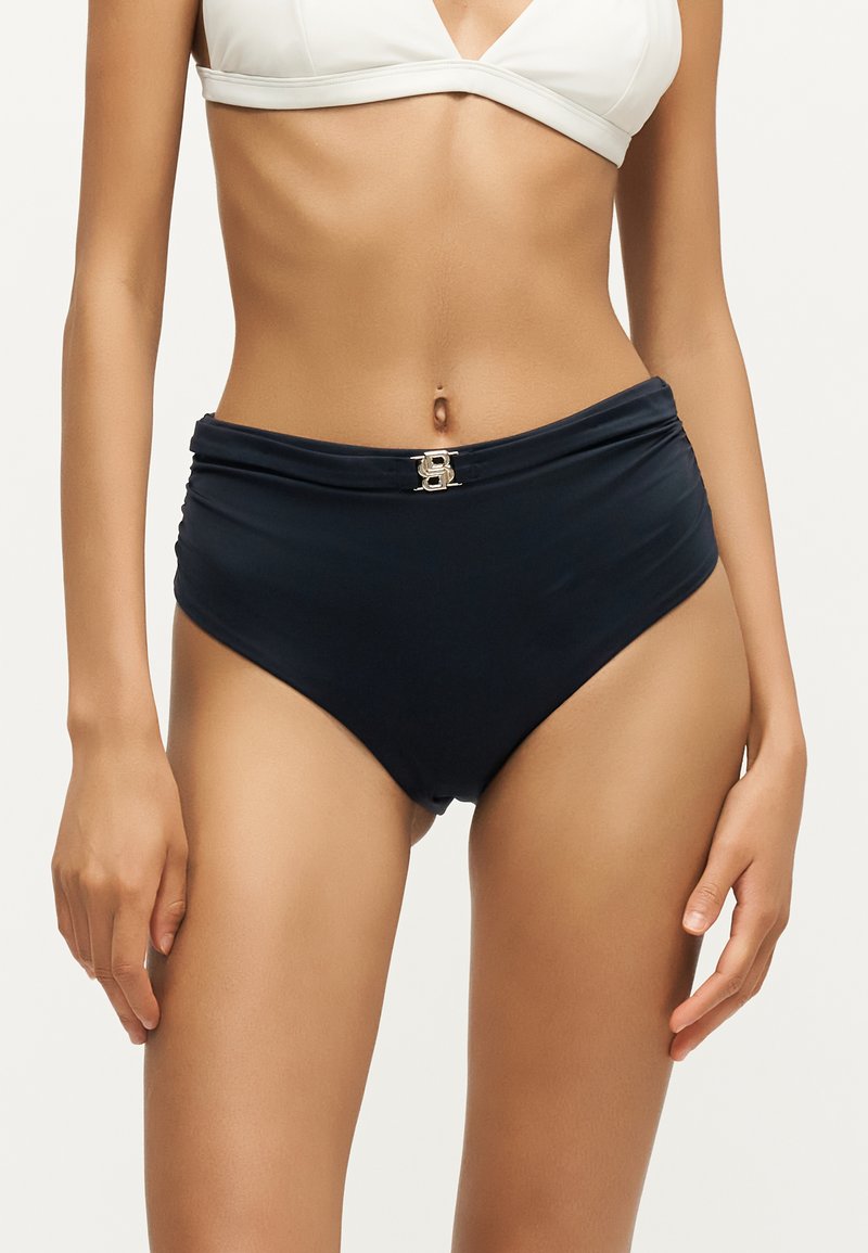 Femme portant un bas de bikini noir taille haute avec un haut de bikini blanc, montrant une boucle décorative dorée sur la ceinture.