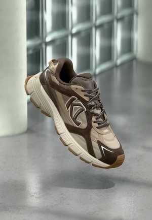 Baskets athlétiques marron et beige avec semelle texturée et fermeture à lacets, suspendues en l'air devant un mur de verre moderne flou en arrière-plan.