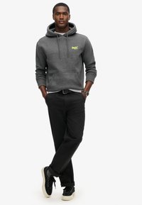 Felpa grigia con cappuccio e tasca frontale, dotata di un piccolo logo neon. Abbinata a pantaloni neri e sneakers nere, con suole bianche.