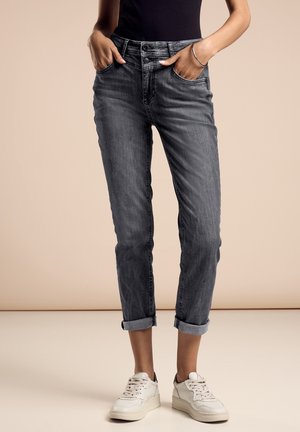 Osoba w podwiniętych ciemnoszarych dżinsach, białych trampkach, czarnej bluzce, z rękami w kieszeniach, na neutralnym tle.