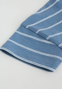 Modrý látkový rukáv s horizontálnymi bielymi pruhmi, zobrazujúci detailné stehy na manžete na svetlom pozadí.