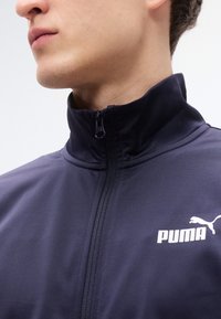 Μπλε ναυτικό μπουφάν με φερμουάρ, με ψηλό γιακά, κατασκευασμένο από λείο ύφασμα, με λευκό λογότυπο Puma στο στήθος και μαύρο φερμουάρ.