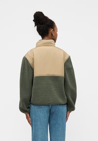 Φλις μπουφάν με μπεζ πάνω μέρος, πράσινο κάτω τμήμα και γιακά. Διαθέτει άνετη εφαρμογή και ριμπ μανσέτες. Ελαφρύ και υφασμένο υλικό.