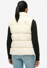 Crème puffer vest met gequiltte delen en hoge kraag. Zwarte kabelgebreide mouwen en logo-opdruk op de bovenrug. Licht denim jeans.