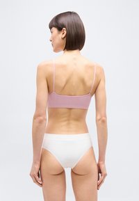 Γυναίκα με κοντά καστανά μαλλιά στέκεται με την πλάτη στραμμένη, φοράει ένα ανοιχτό ροζ μπραλέ και λευκά αόρατα εσώρουχα μπροστά σε ένα απλό φόντο.