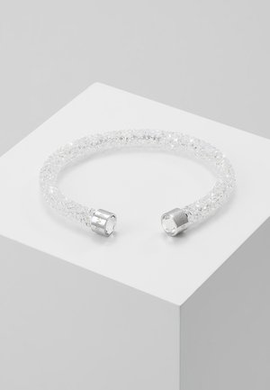 Open armband bezet met kristallen en zilverkleurige metalen uiteinden, liggend op een wit geometrisch oppervlak.