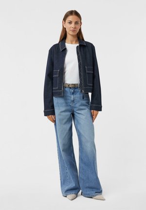 Junge Frau steht mit einer dunklen Jeansjacke, einem weißen Hemd, hellblauen weit geschnittenen Jeans, einem grauen Gürtel und grauen spitzen Schuhen vor einem weißen Hintergrund.