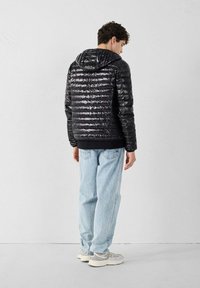 Giubbotto imbottito nero con finitura lucida, cuciture orizzontali e cappuccio; abbinato a jeans azzurri chiari e sneaker in tonalità neutre.