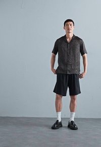 Uomo in piedi contro una parete semplice, indossa una camicia a maniche corte con motivo, pantaloni neri, calze bianche e scarpe eleganti nere.